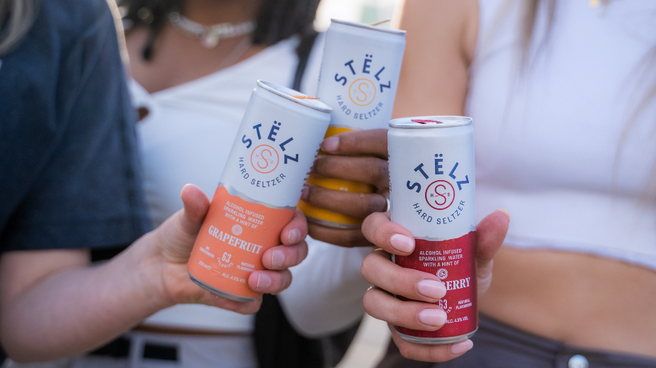 Three hands holding STËLZ Hard Seltzer cans outdoors in the sun
